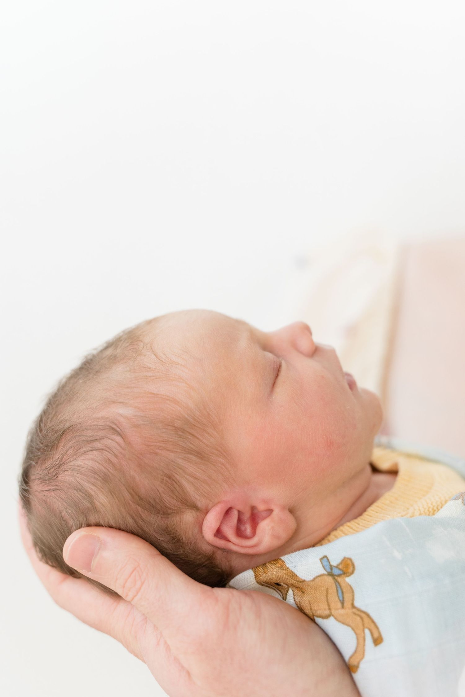 A newborn baby sleeping peacefully while being cradled in an adult's hand, wearing a light blue outfit with a deer print.
