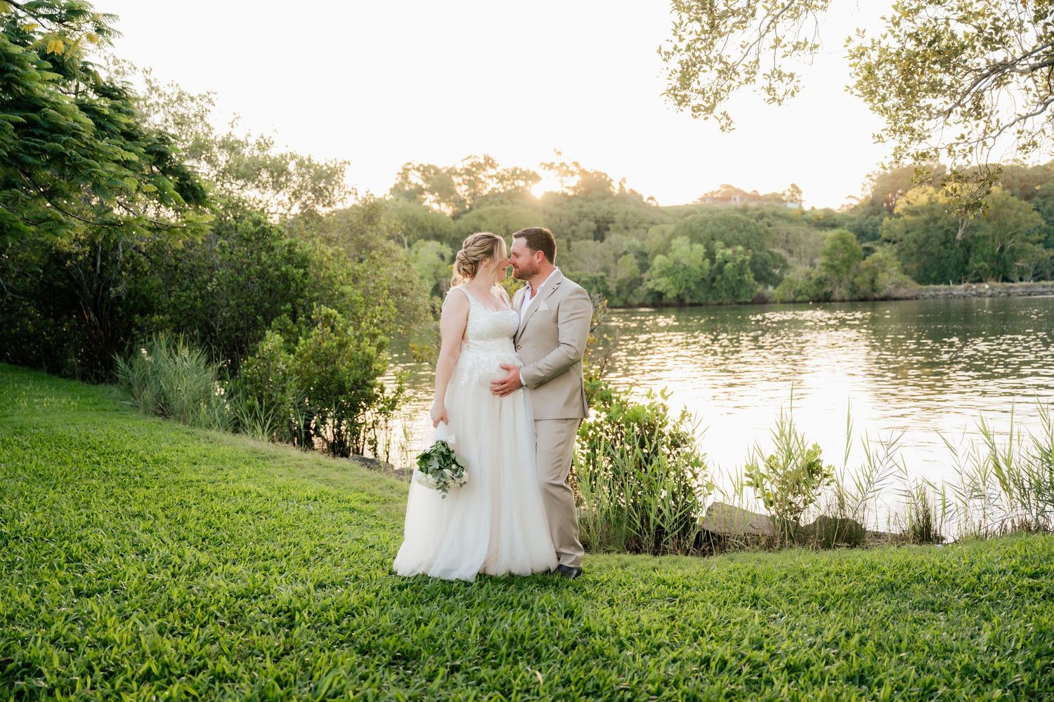 Bride and groom share a kiss by a sunlit pond surrounded by lush greenery on their wedding day.