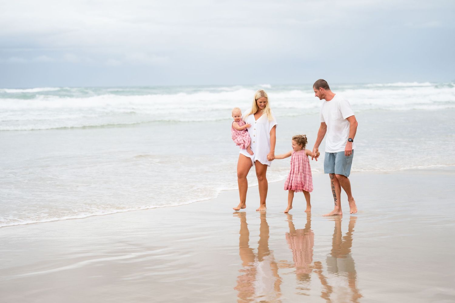 Family of four holding hands walking on a sandy beach near ocean waves on a cloudy day.