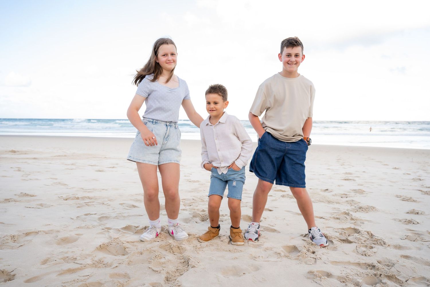Three children posing together on a sandy beach, smiling for a family photo on a cloudy day.
