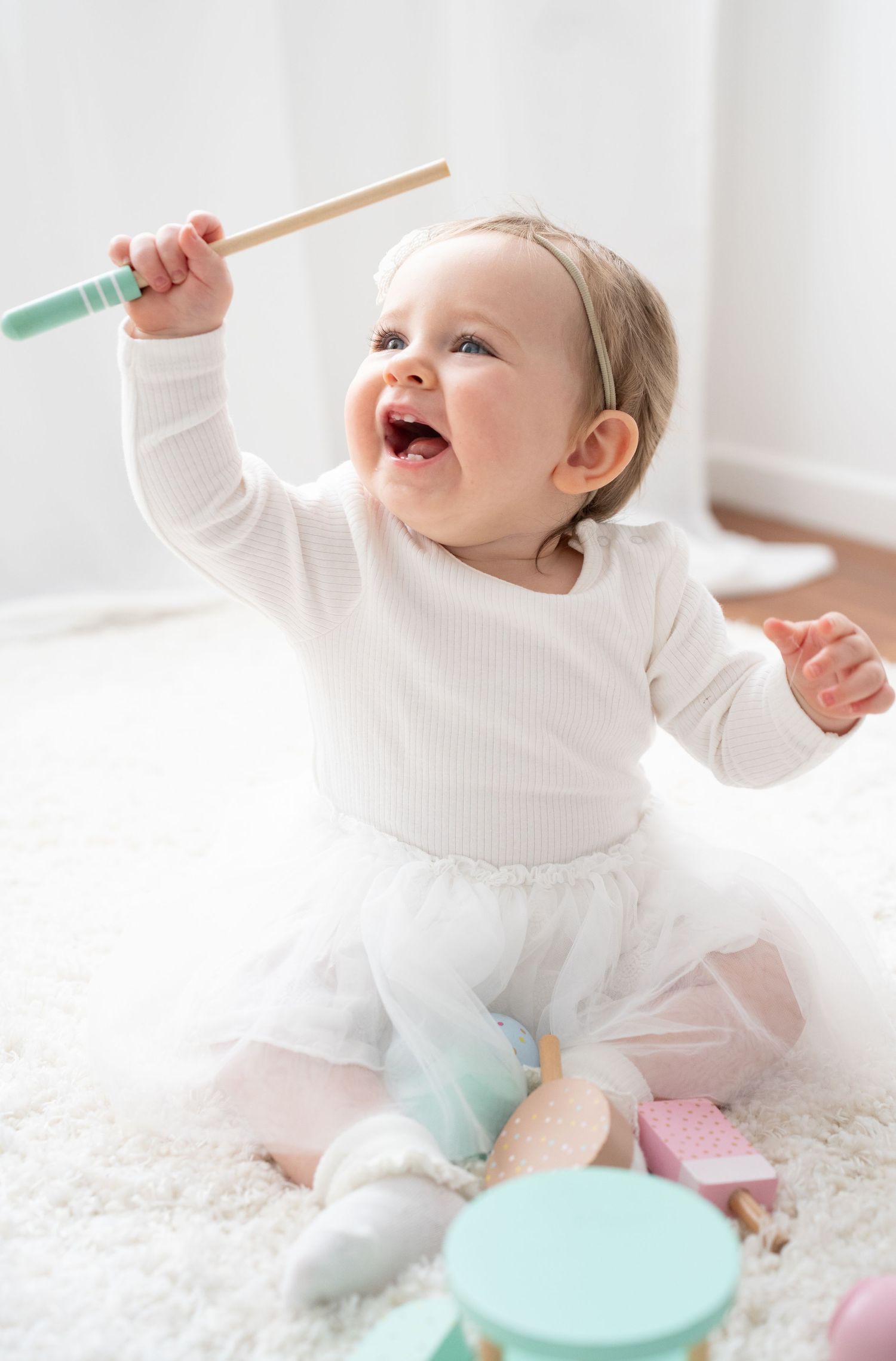 Happy baby girl in white tutu dress playing with wooden drum sticks, sitting on soft carpet with colorful toys.