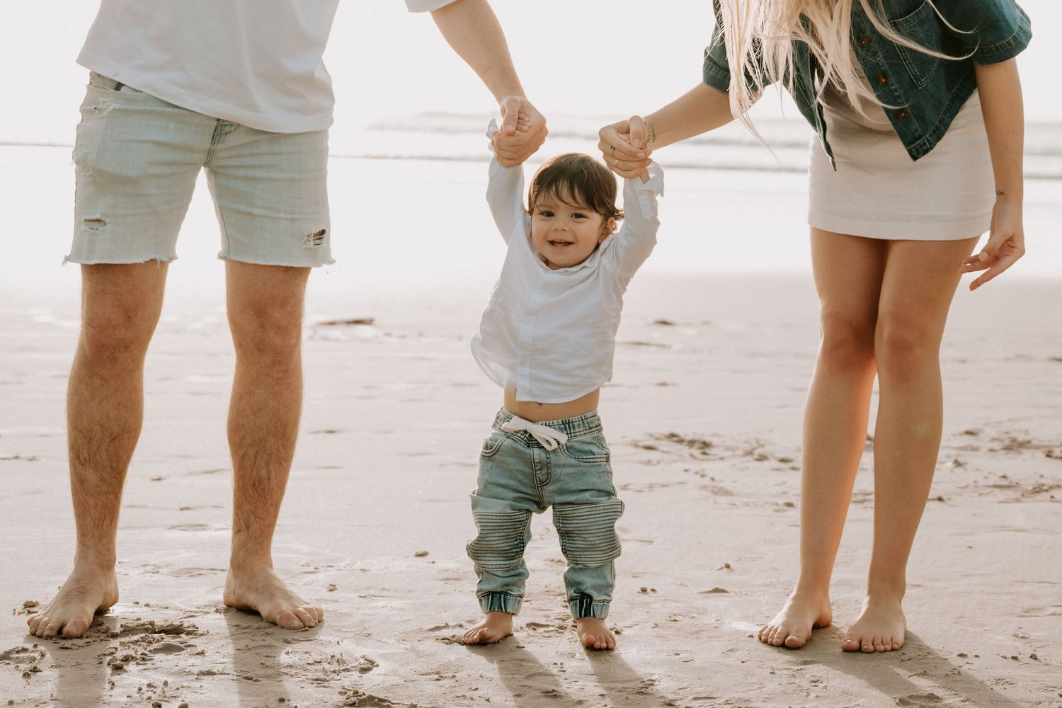 Toddler taking first steps on sandy beach held by parents hands, family beach photo session.
