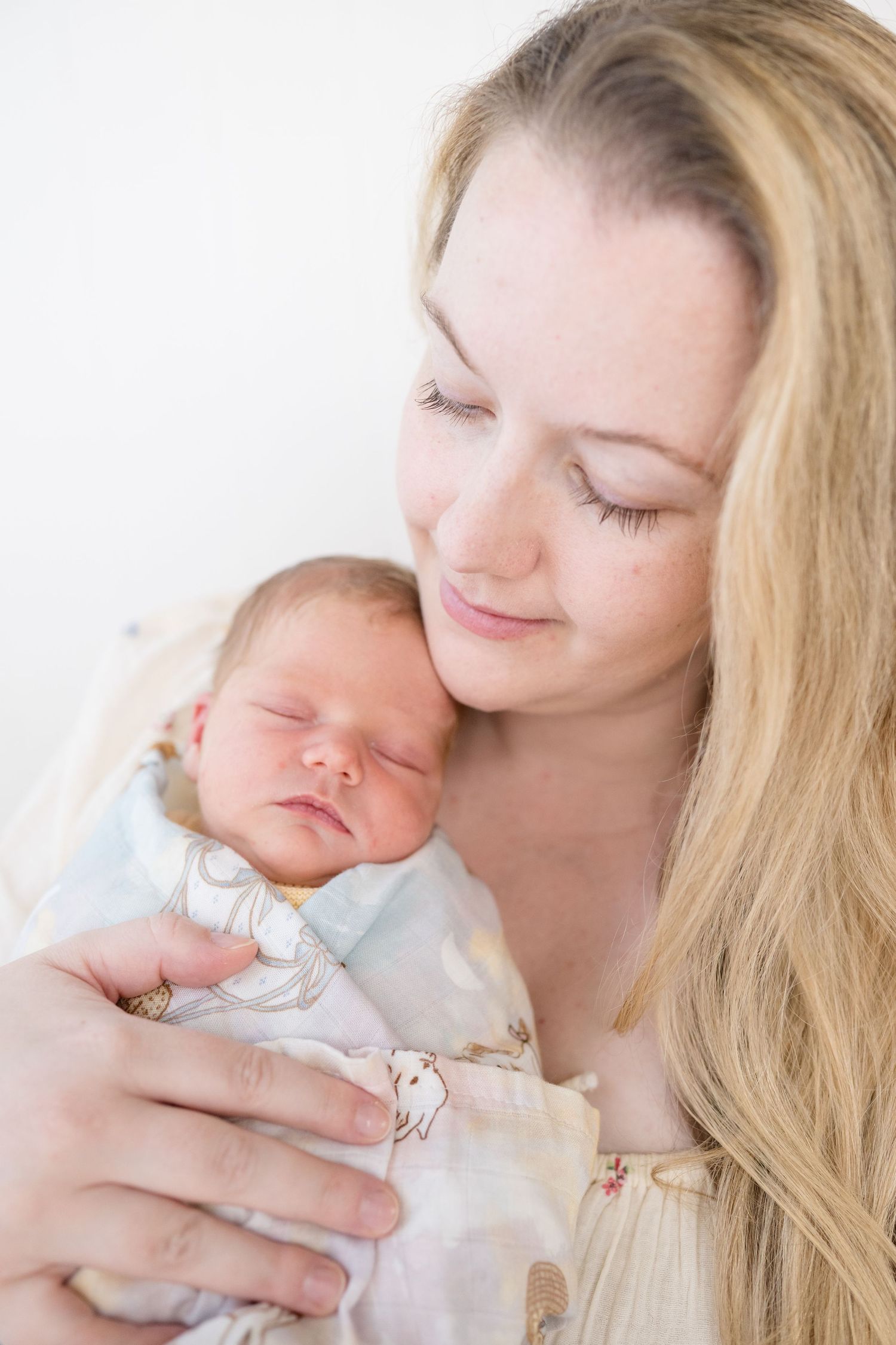 A young blonde mother tenderly holds her sleeping newborn baby wrapped in a soft blanket.