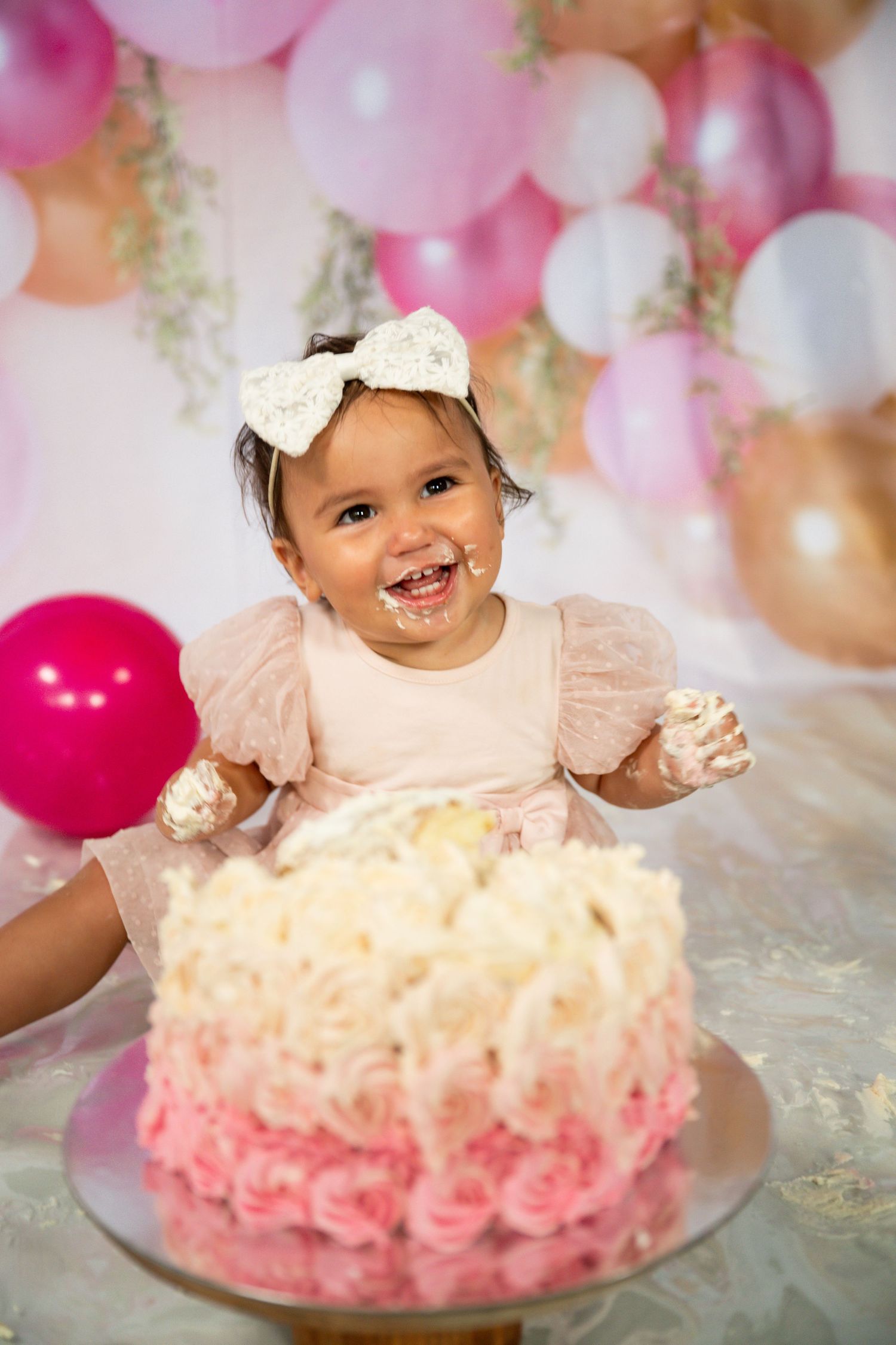 A baby in a pink tulle dress sits among pink and gold balloons during a cake smash photo session.