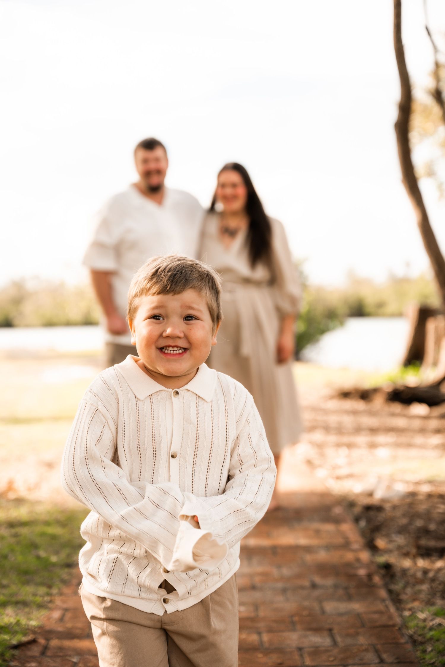 A young child in a white striped shirt and khaki pants smiles joyfully on a brick path while family stands in background.