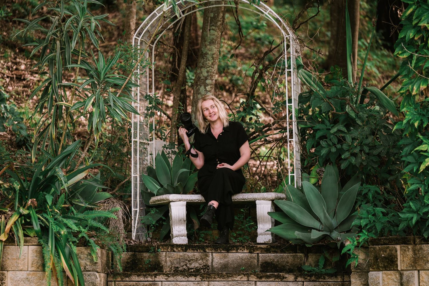 A couple embraces in a lush garden setting with stone walls and tropical plants creating a romantic atmosphere.