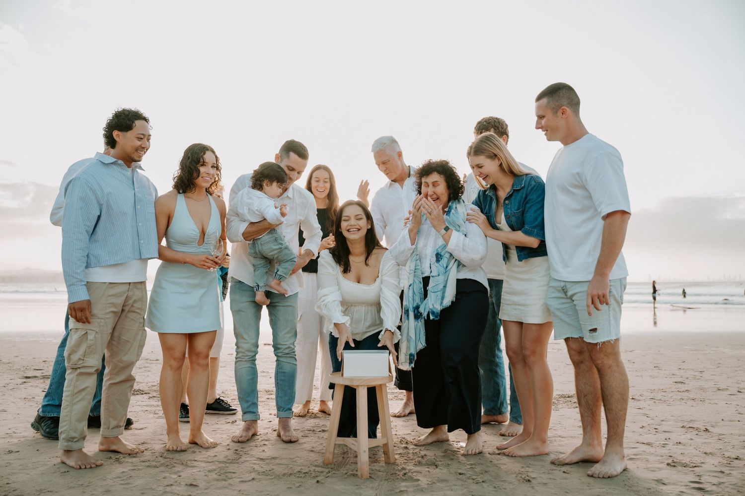 Large family group smiling and celebrating together on a sandy beach at sunset in casual summer outfits.