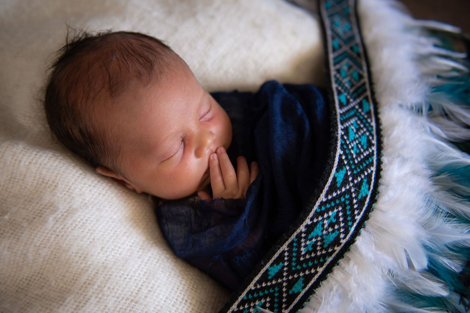 Newborn baby swaddled in navy blue wrap sleeping peacefully in a white fluffy basket with decorative beaded trim.