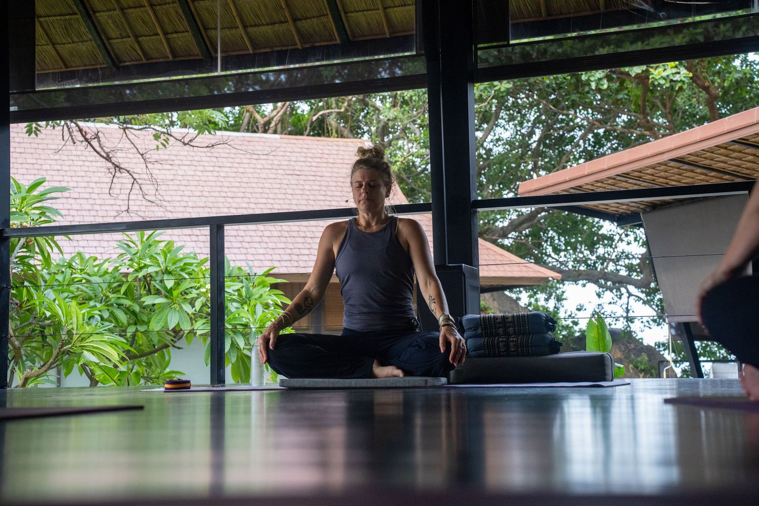 Woman meditating in lotus pose on yoga mat in open-air studio surrounded by tropical greenery.