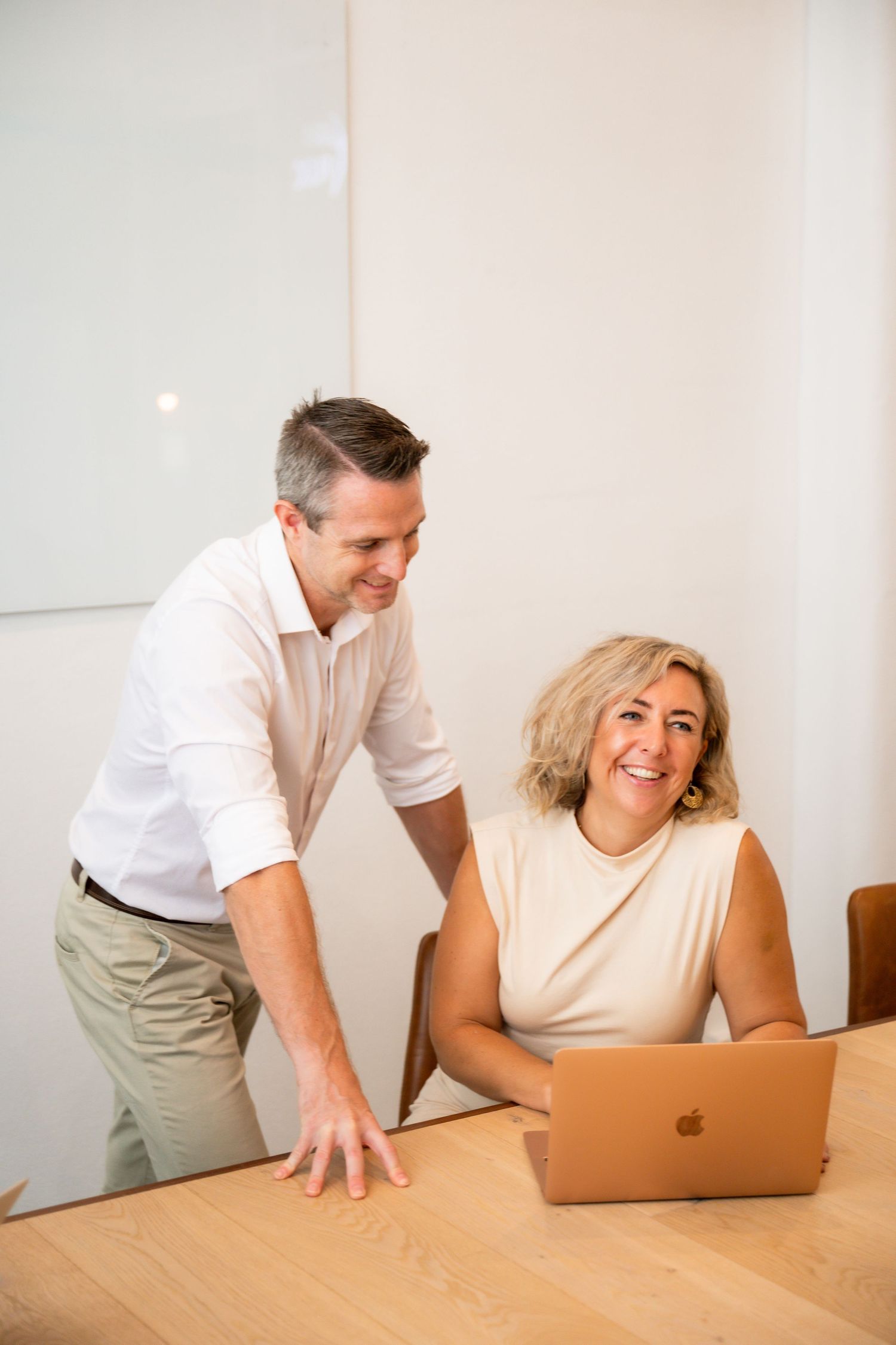 Two coworkers collaborate at a wooden desk while working on a MacBook laptop in a bright office space.