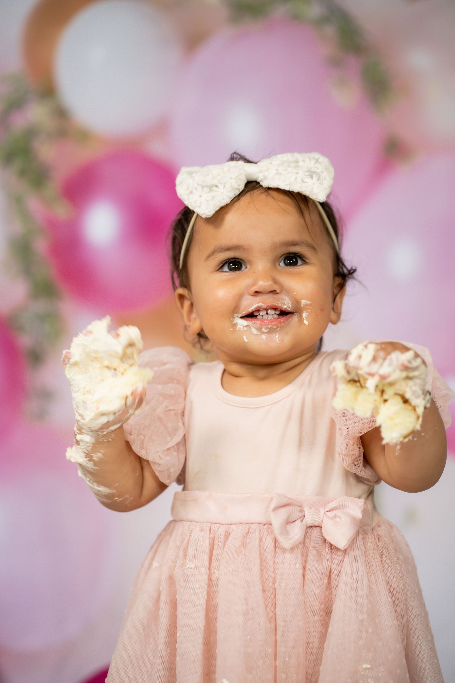 Baby wearing pink polka dot dress and white bow smiling during a portrait session with pink floral backdrop.