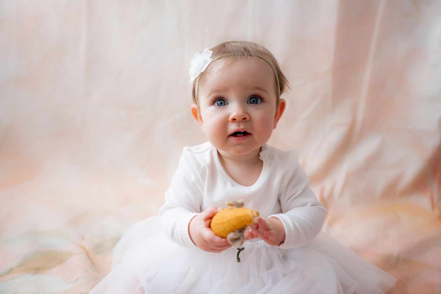 A cute baby girl in a white dress and bow holds a yellow lemon while sitting against a soft pink background.