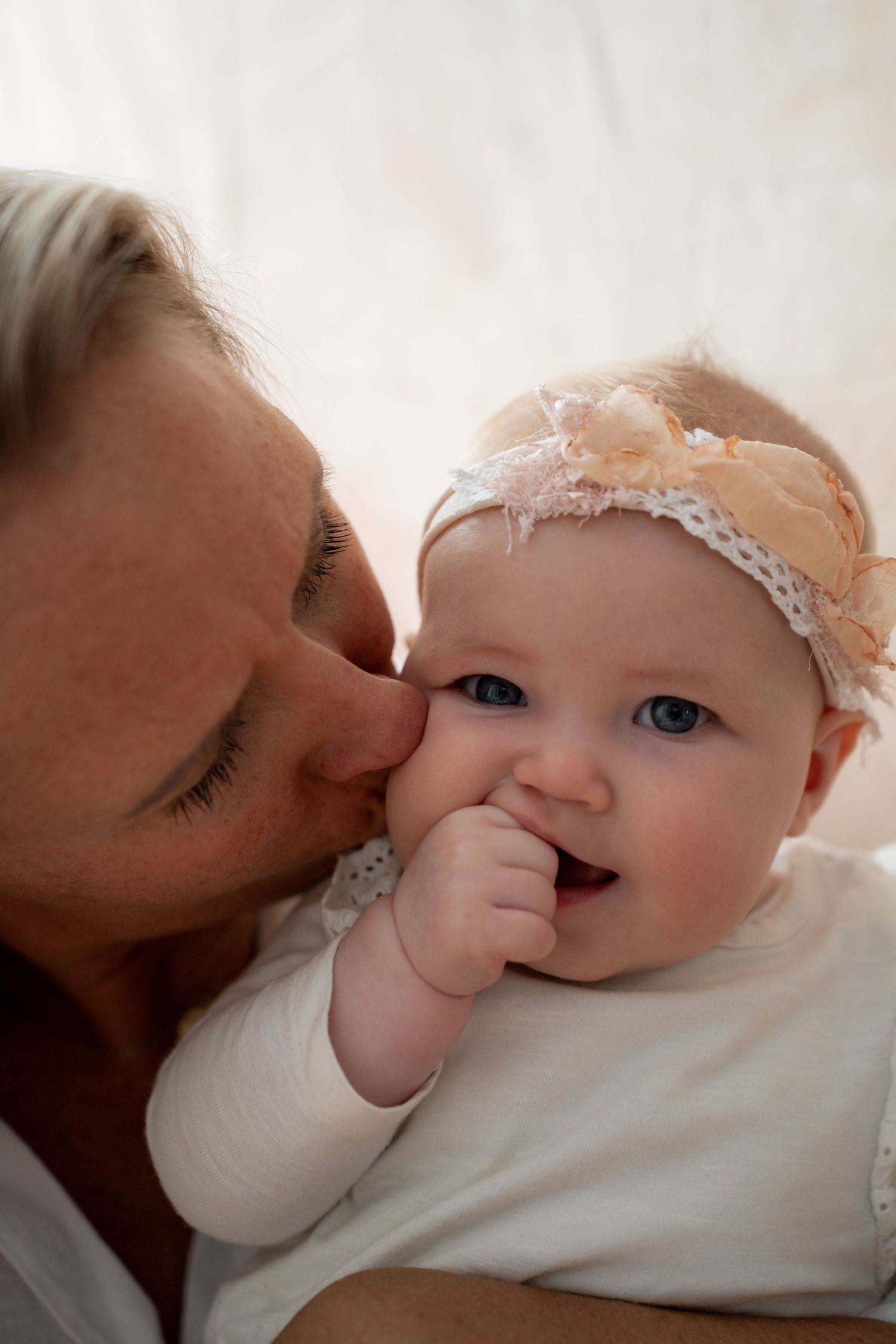 A mother tenderly kisses her smiling blue-eyed baby girl wearing a peach floral headband.