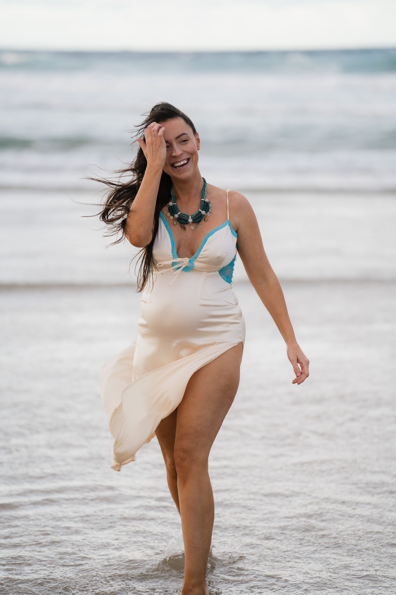Smiling woman in cream wrap dress and turquoise bikini top walks along ocean shoreline with wind blowing her dark hair.