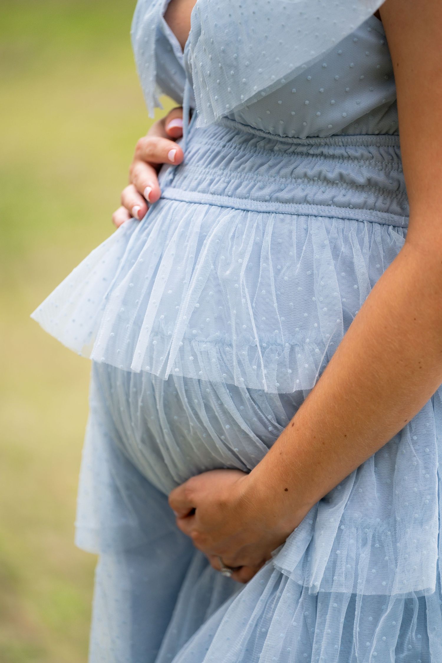 Pregnant woman in a light blue dotted tulle dress gently cradling her baby bump outdoors.