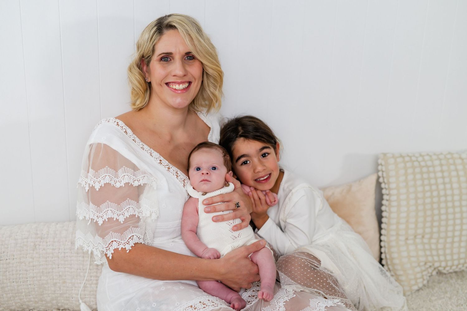 A smiling woman in white lace holds a newborn baby while an older child leans in beside them on a white couch.