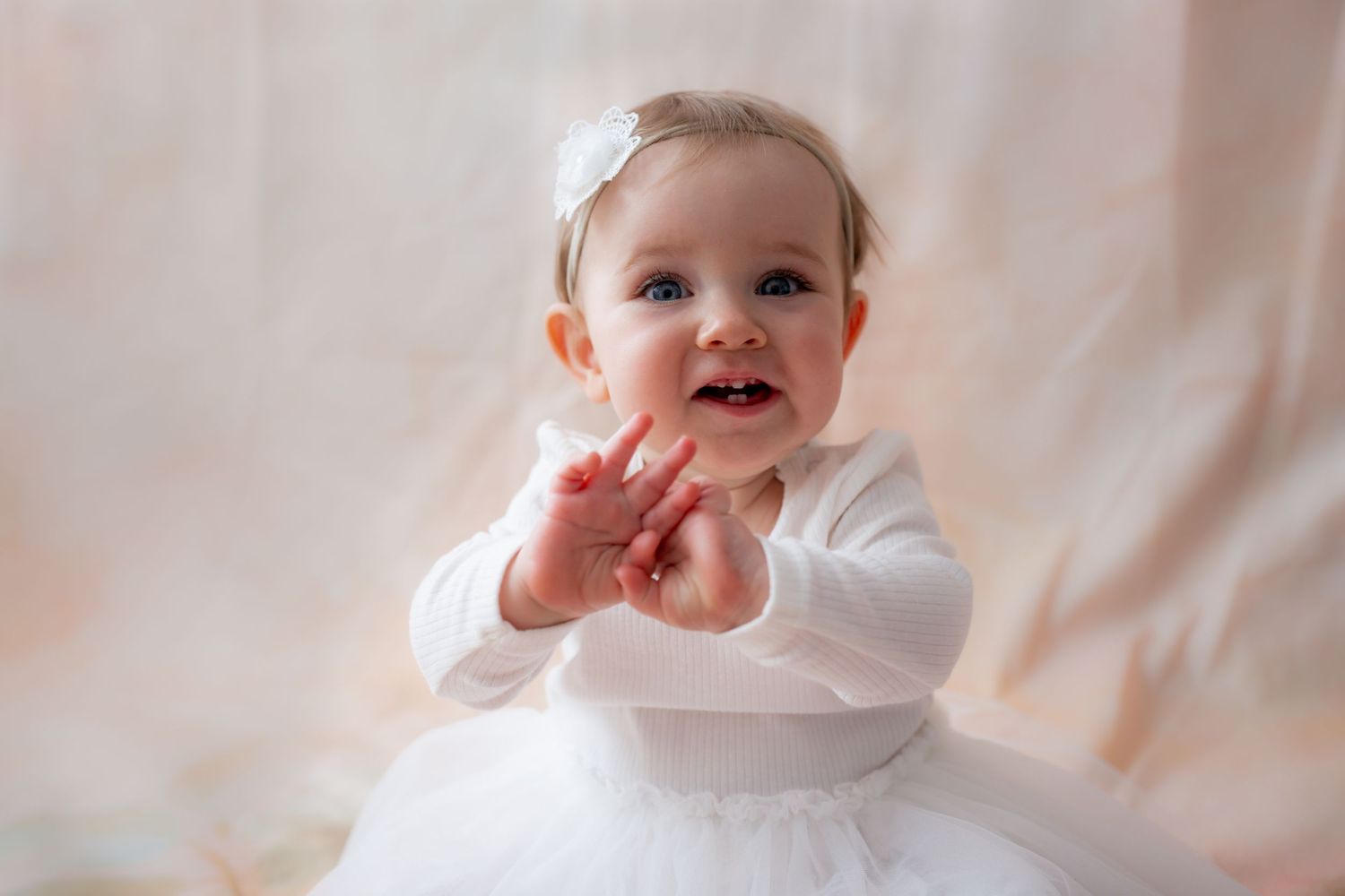 Adorable baby girl in white dress and headband clapping hands, smiling joyfully against soft pink background.