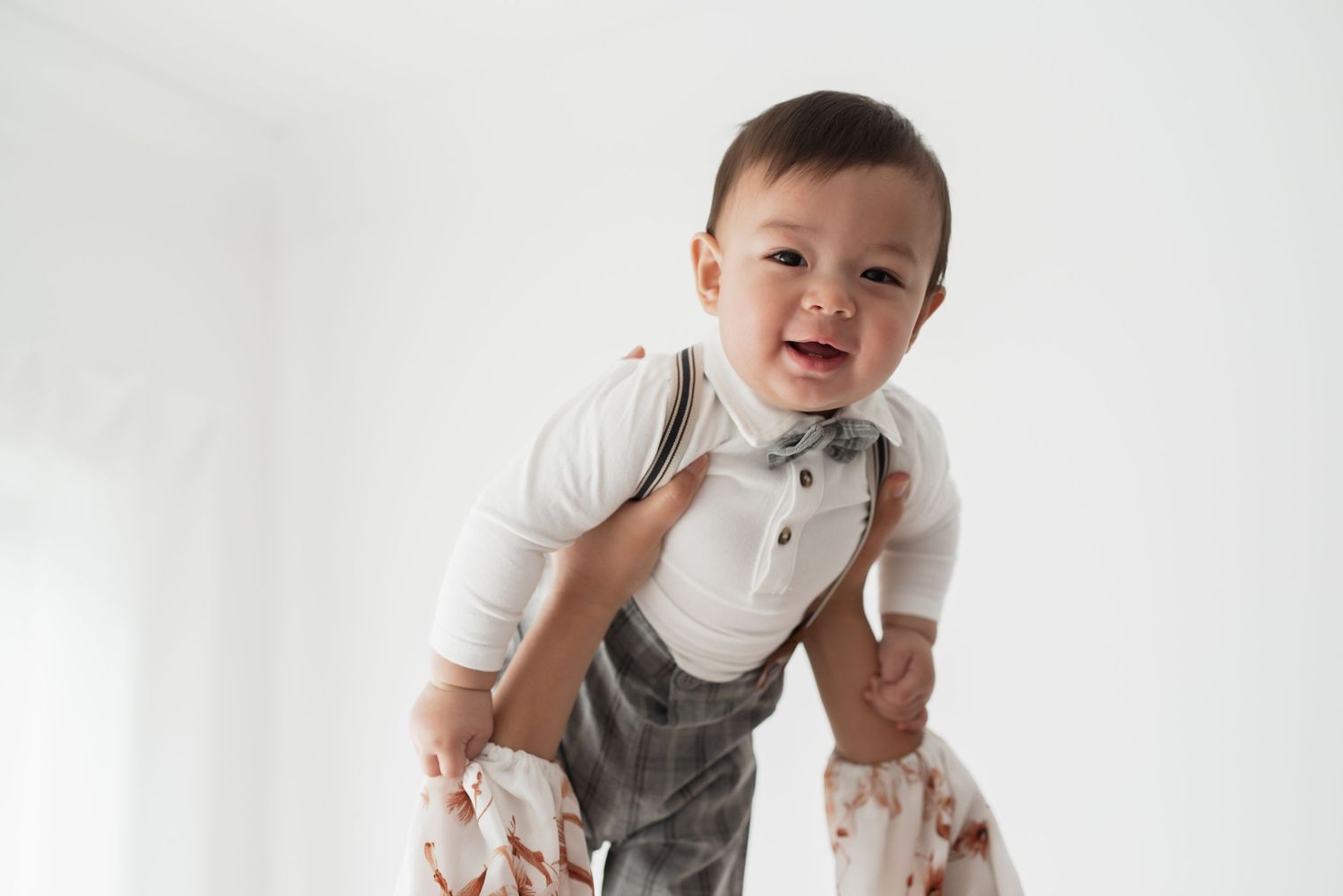 Smiling baby boy in white onesie with bow tie and suspenders being held up against a white background.