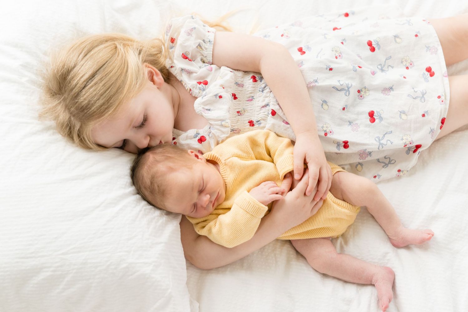 Young girl in floral dress gently kisses sleeping newborn baby sibling lying on white bed.