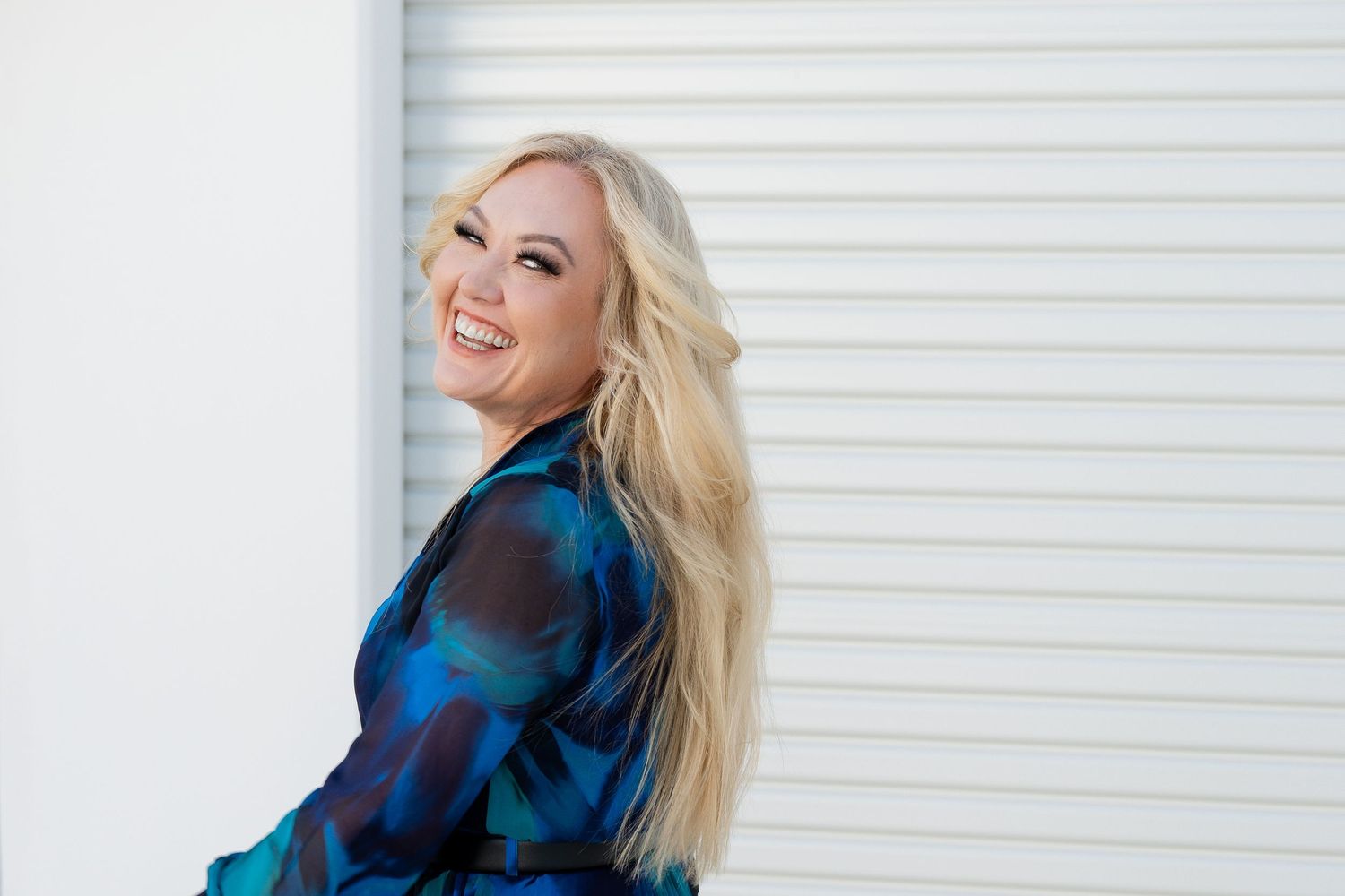 Smiling blonde woman in a blue patterned blouse posing against a white wall, looking over her shoulder.