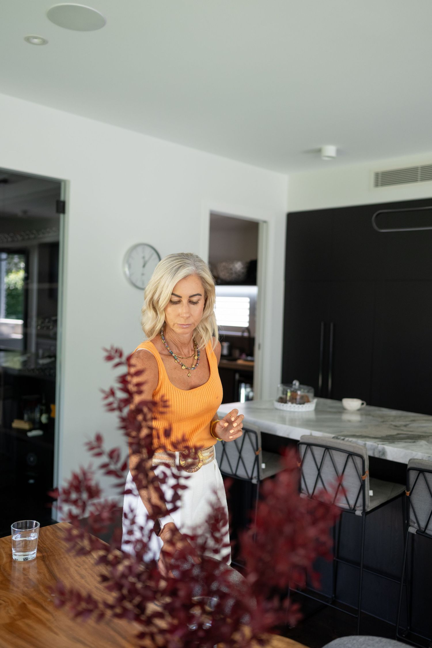 Blonde woman in orange top arranges red floral stems on wooden table in modern kitchen with marble island.