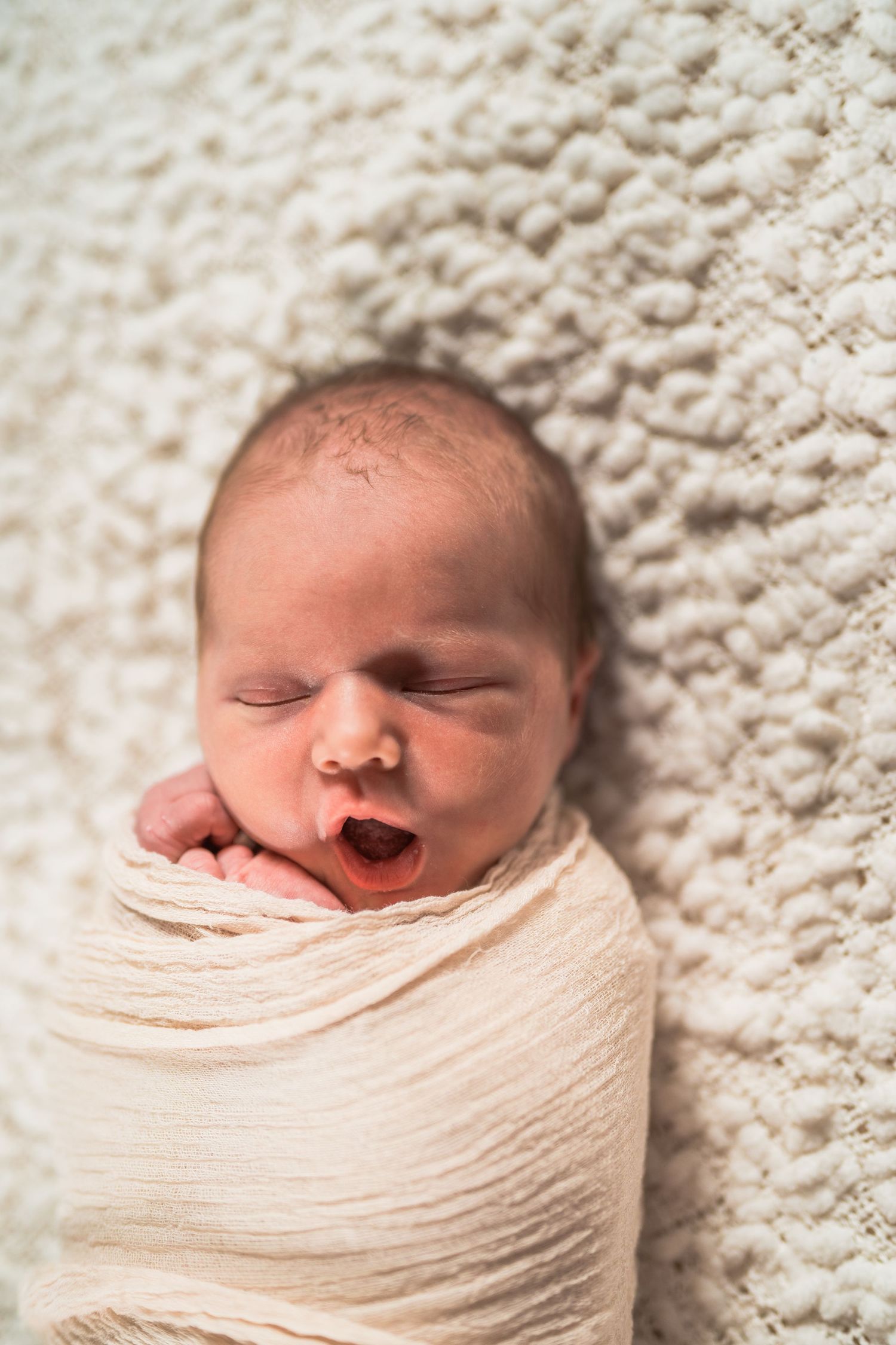 Newborn baby swaddled in white wrap yawning while lying on a soft cream textured blanket.