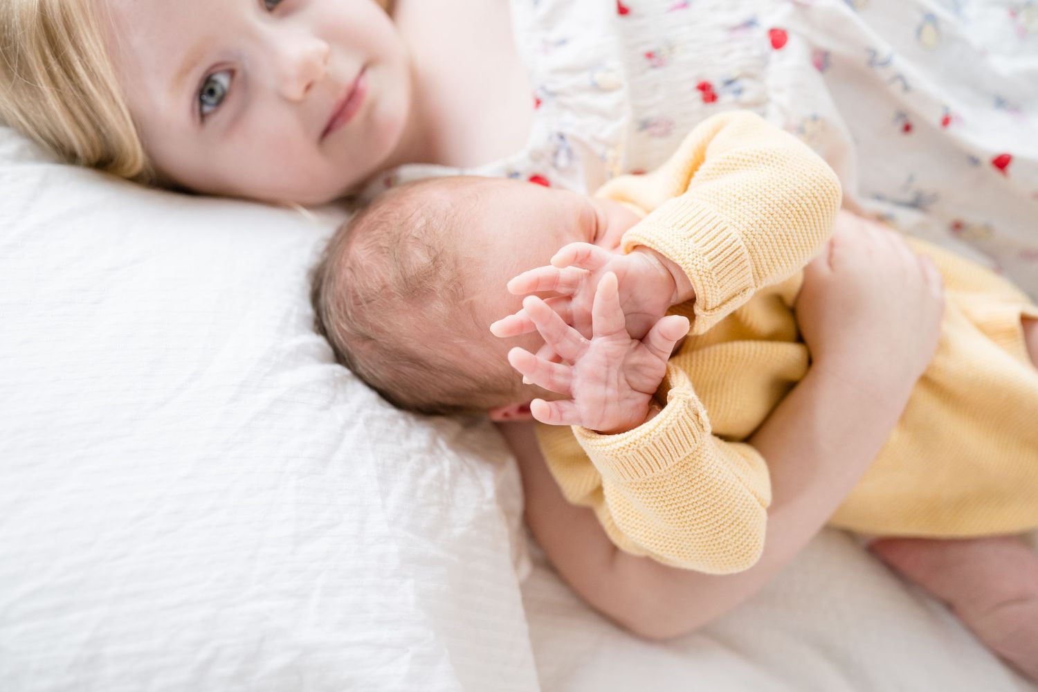 Young girl lying on white bed gently holding a sleeping newborn baby wrapped in a yellow blanket.