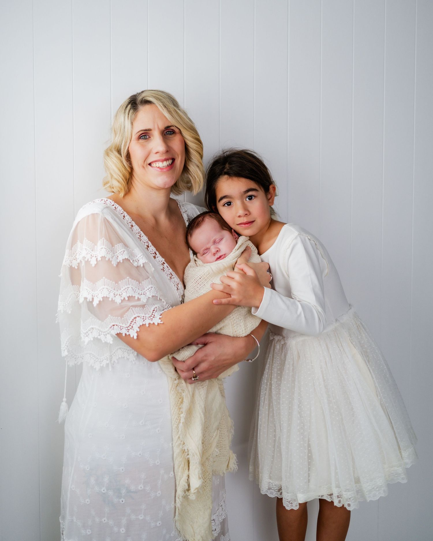 A smiling blonde mother in a white lace dress holds a newborn baby alongside her young daughter in a white dress.