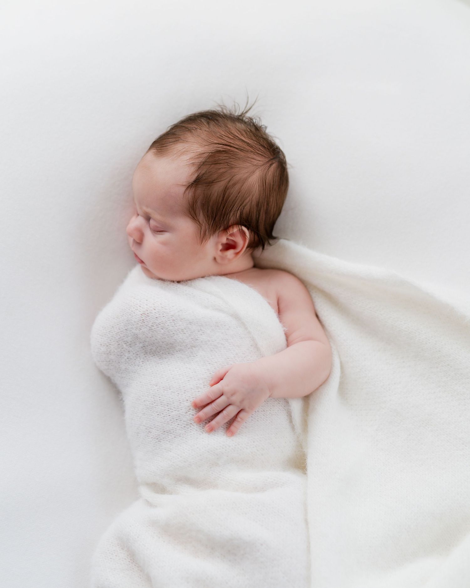 Newborn baby sleeping peacefully wrapped in a soft white blanket on a white background.