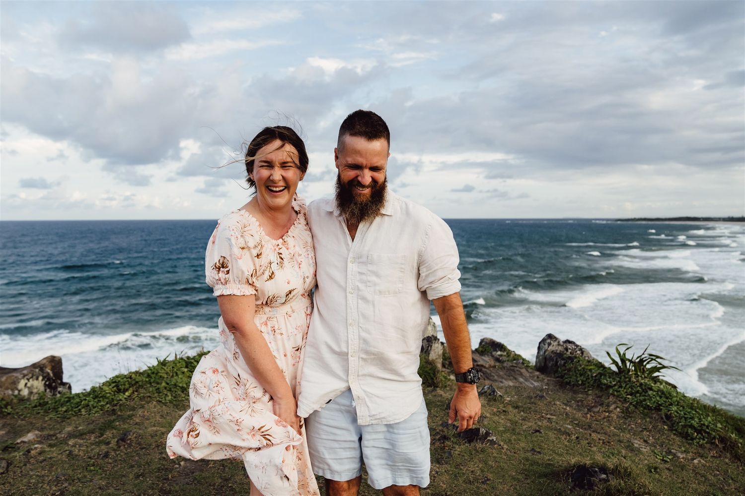 A couple shares romantic moments along a scenic coastal cliff with crashing ocean waves in the background.