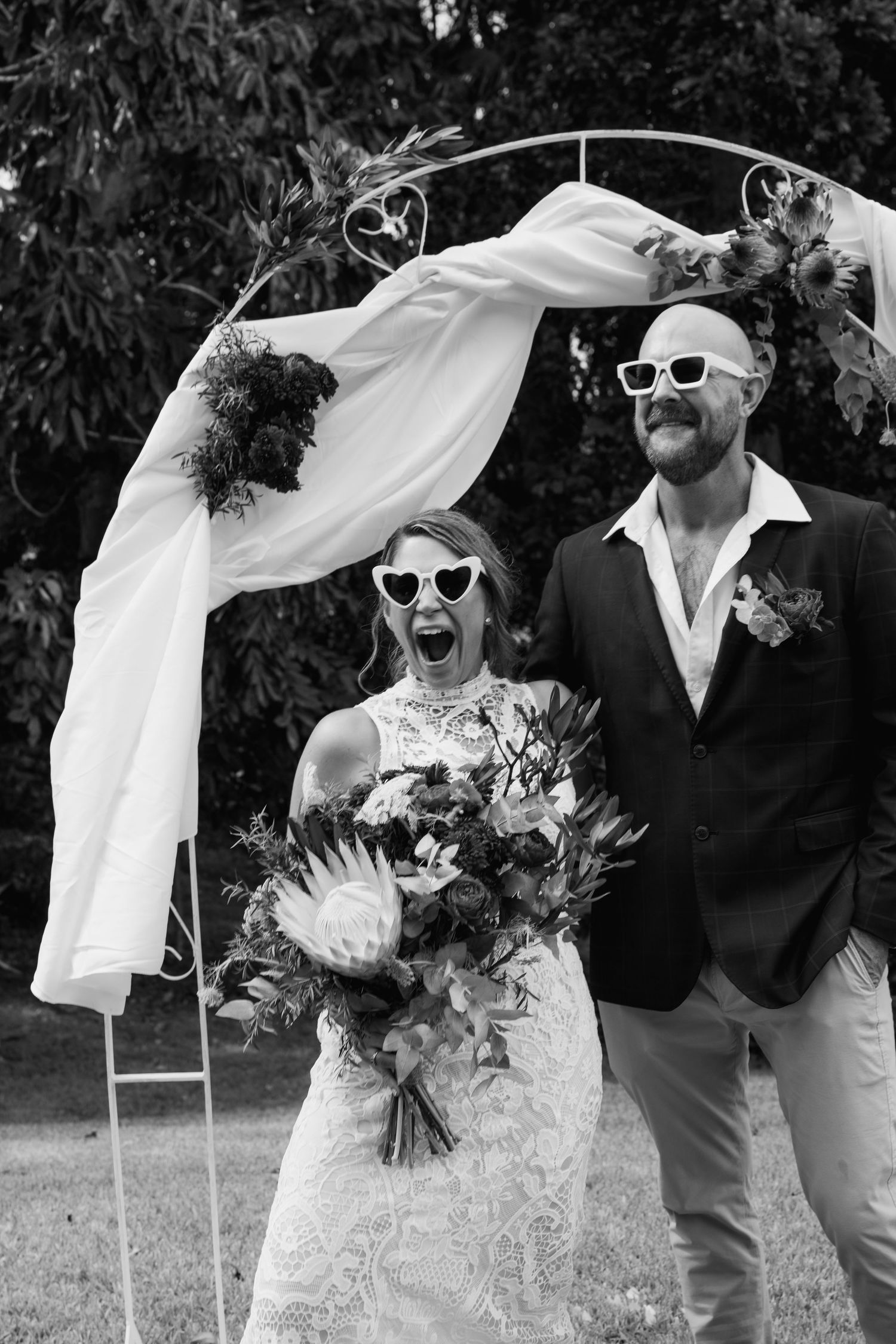 A couple wearing sunglasses poses under an arch at their outdoor wedding ceremony with a flowing veil and bouquet.
