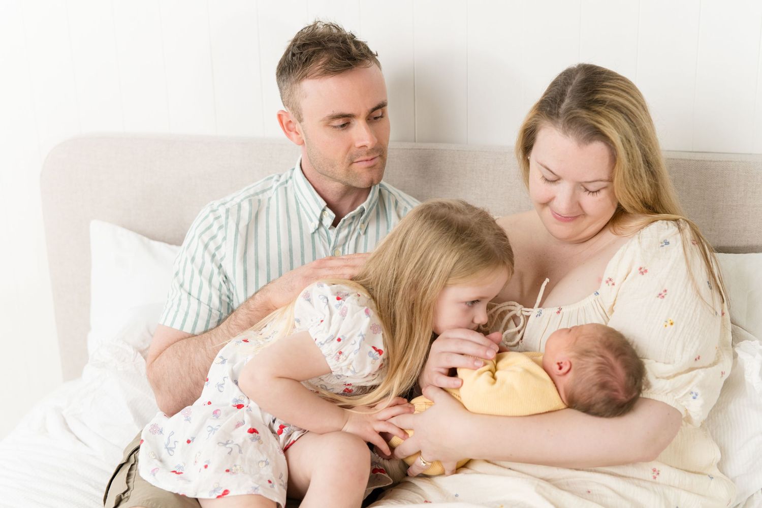Family of four sitting on bed, parents smiling at newborn baby while young daughter lovingly kisses sibling.