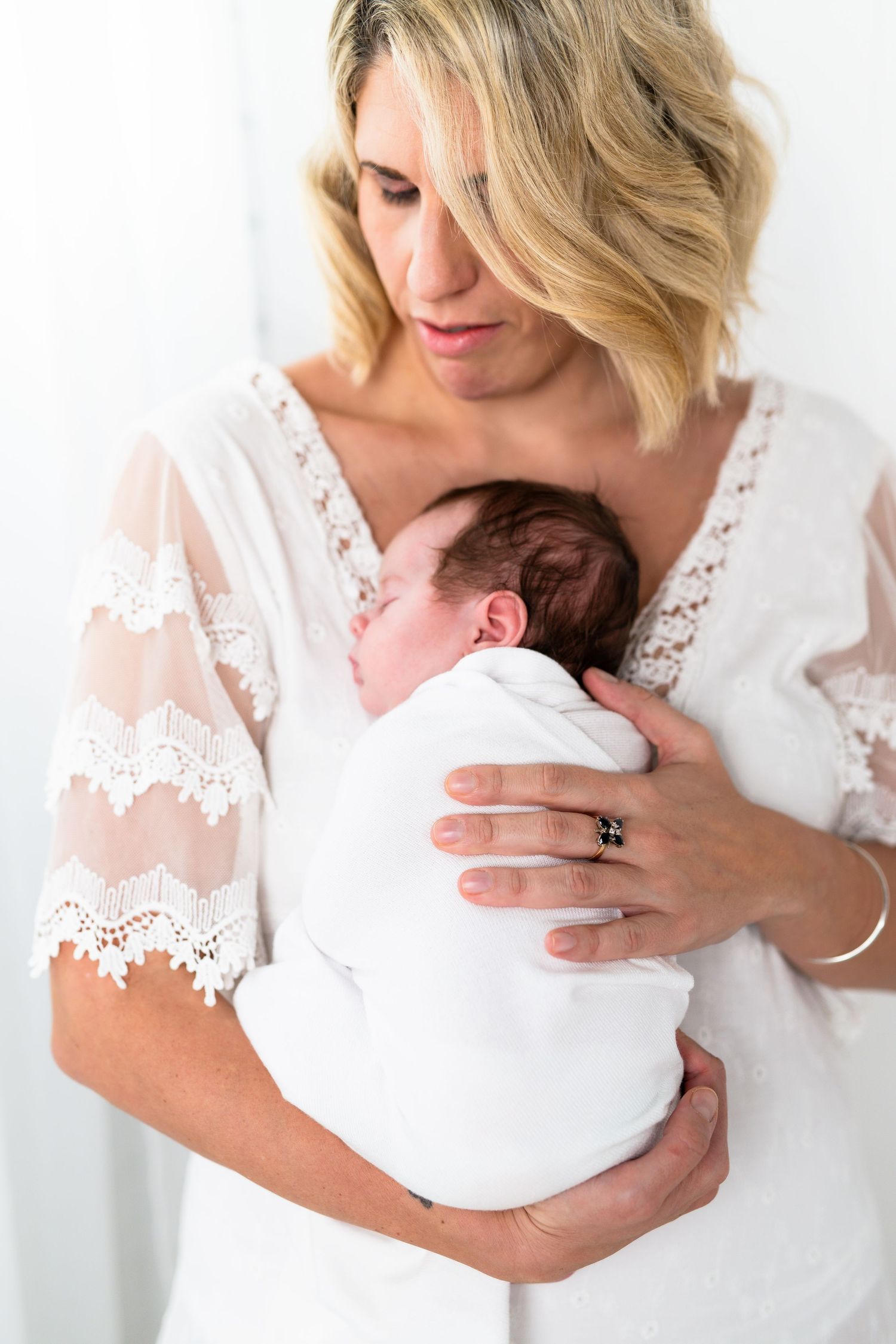 A blonde mother in a white lace dress tenderly holds her sleeping newborn baby wrapped in a white swaddle.