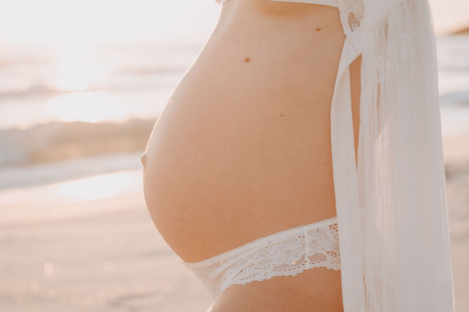 Pregnant woman in white lace underwear and sheer robe showing baby bump at beach during golden hour.