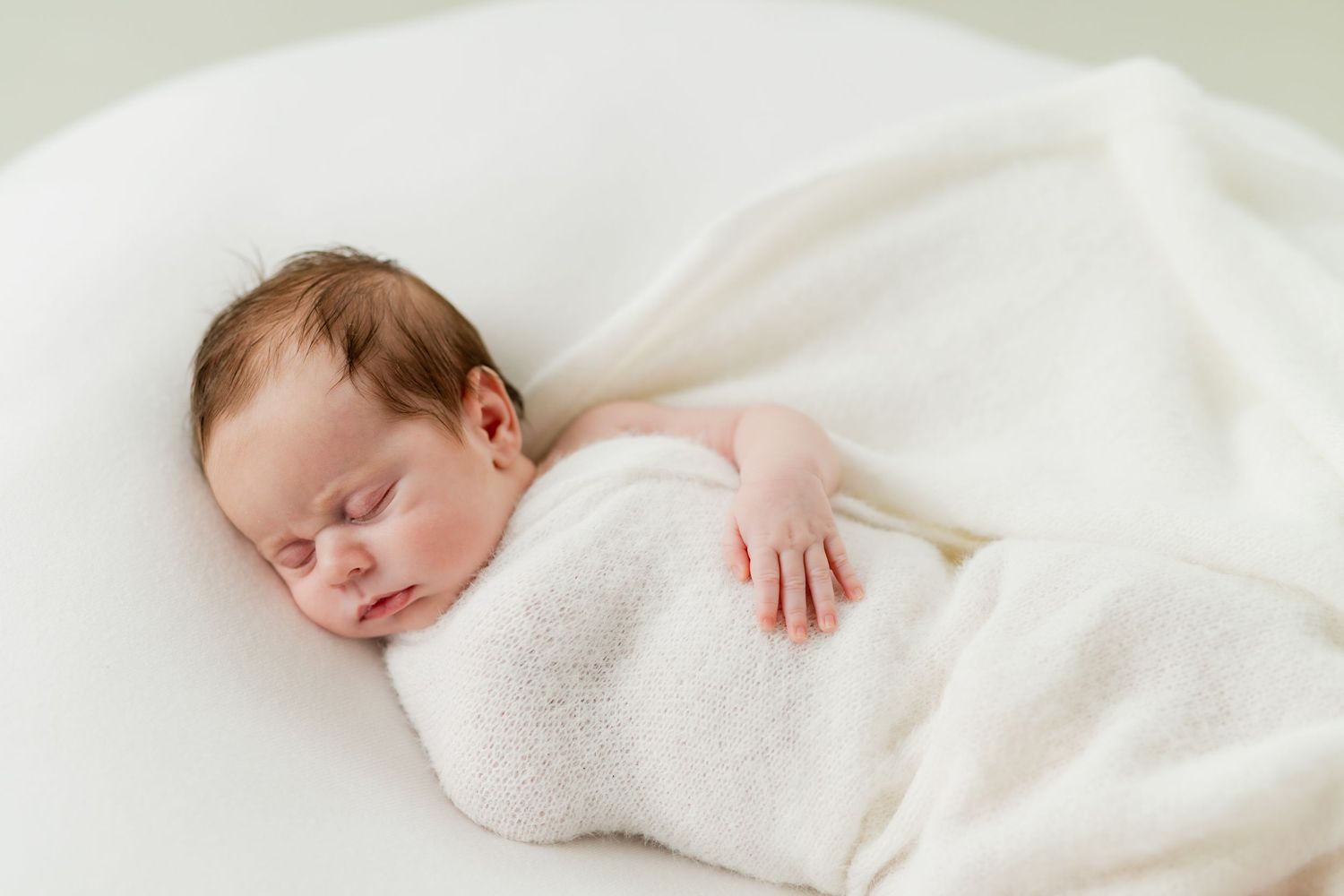 Peaceful newborn baby swaddled in white blanket sleeping on white cushion in soft natural light.
