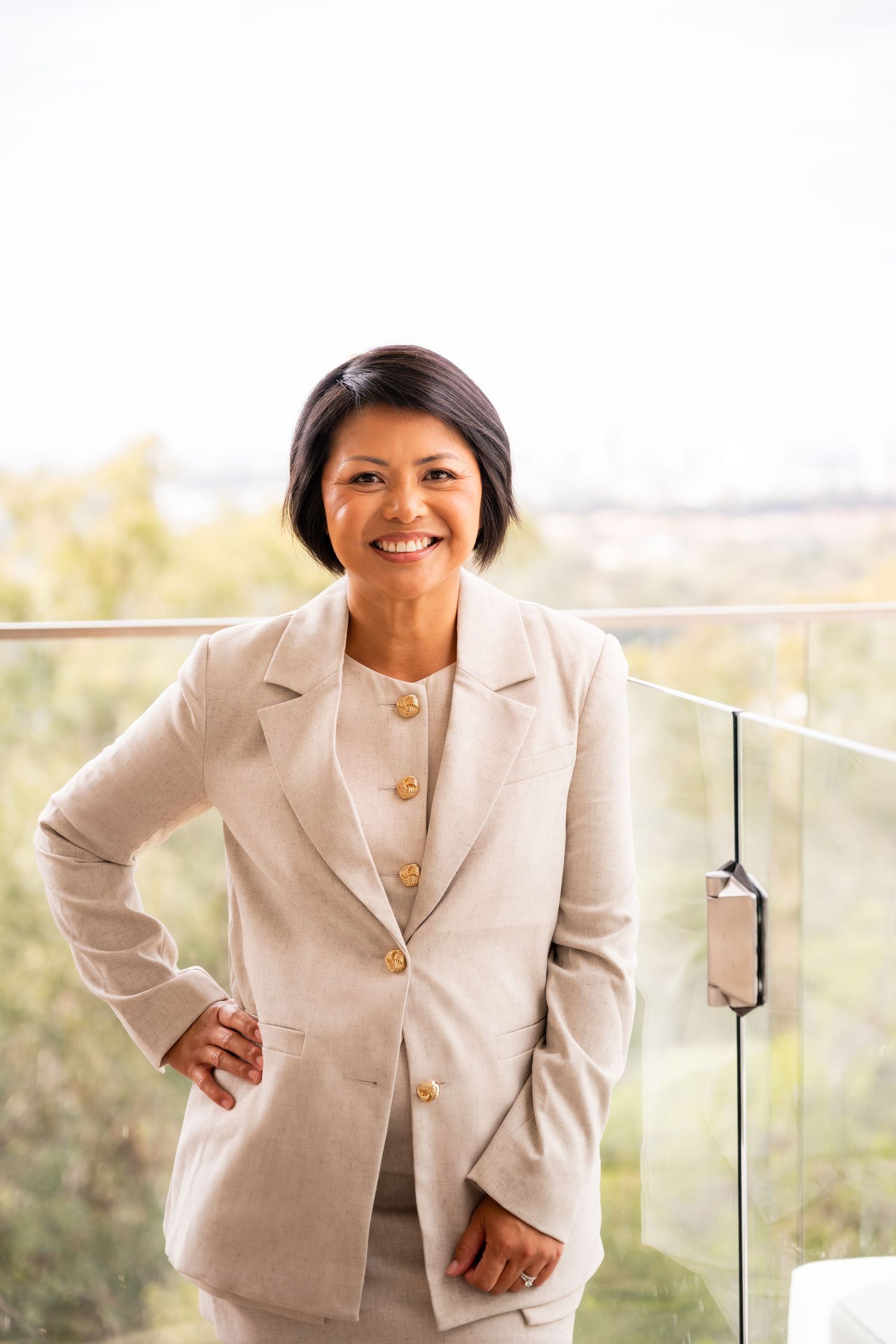 Professional businesswoman in beige suit smiling confidently on modern balcony with scenic outdoor view.