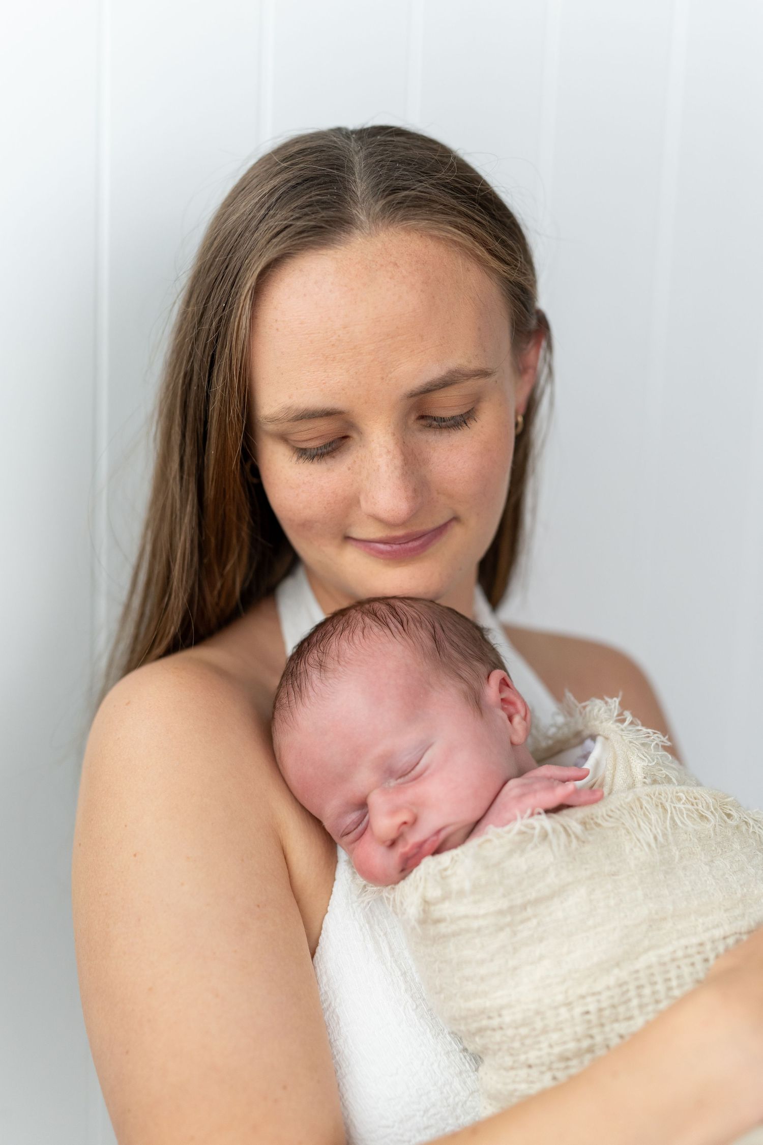 A young mother tenderly holds her sleeping newborn baby wrapped in a soft cream swaddle against a white background.
