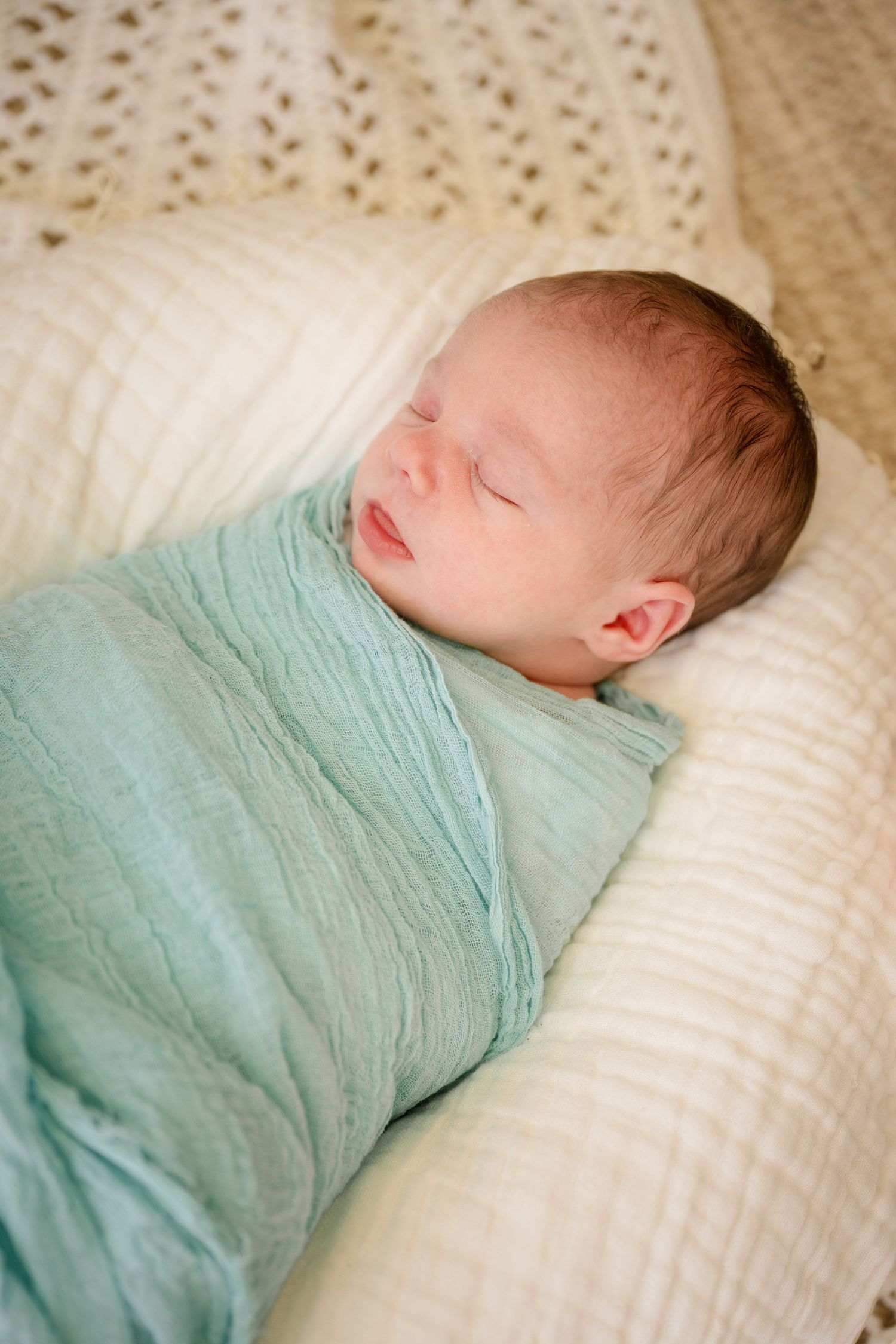 A sleeping baby swaddled in mint green fabric lies peacefully on a white pillow.