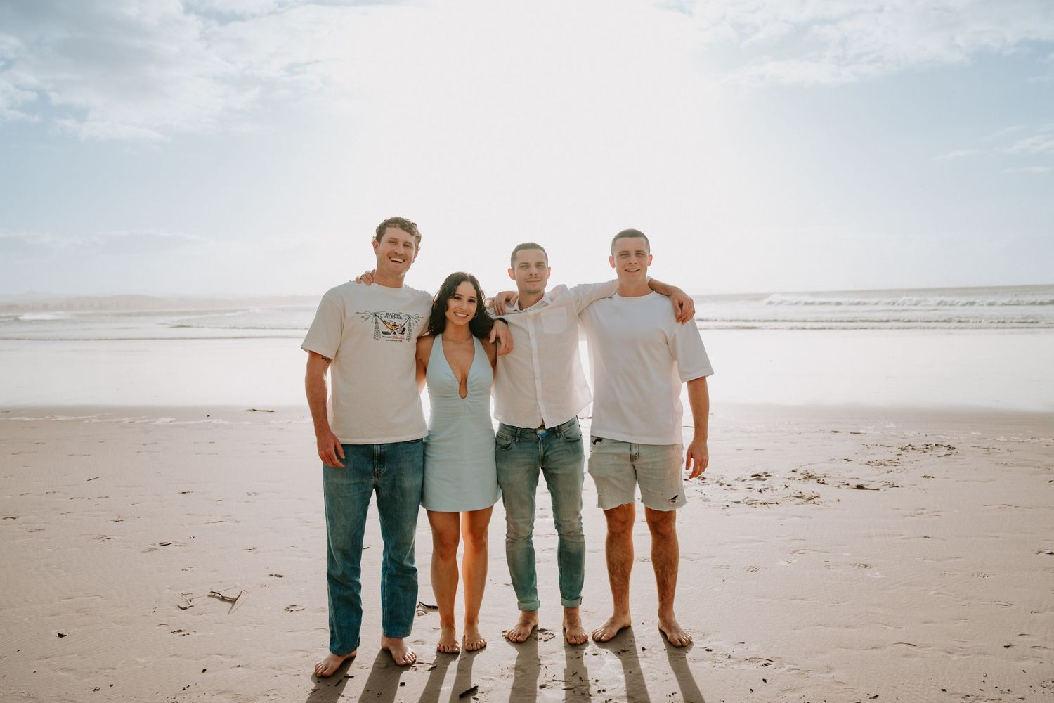 Four friends smiling together on a sandy beach with ocean waves in the background on a sunny day.