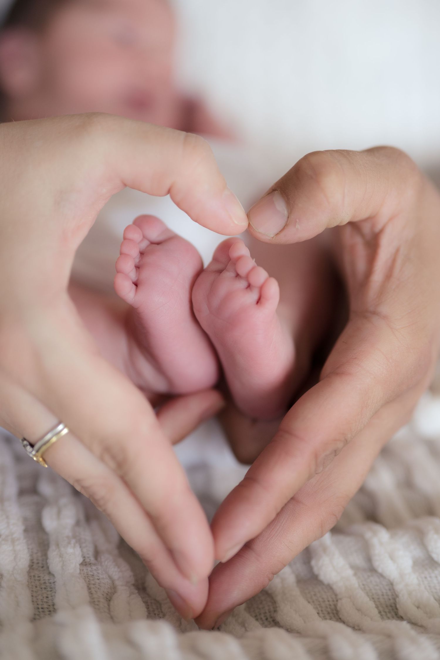 Two parents form a heart shape with their hands around their newborn baby's tiny feet on a soft blanket.