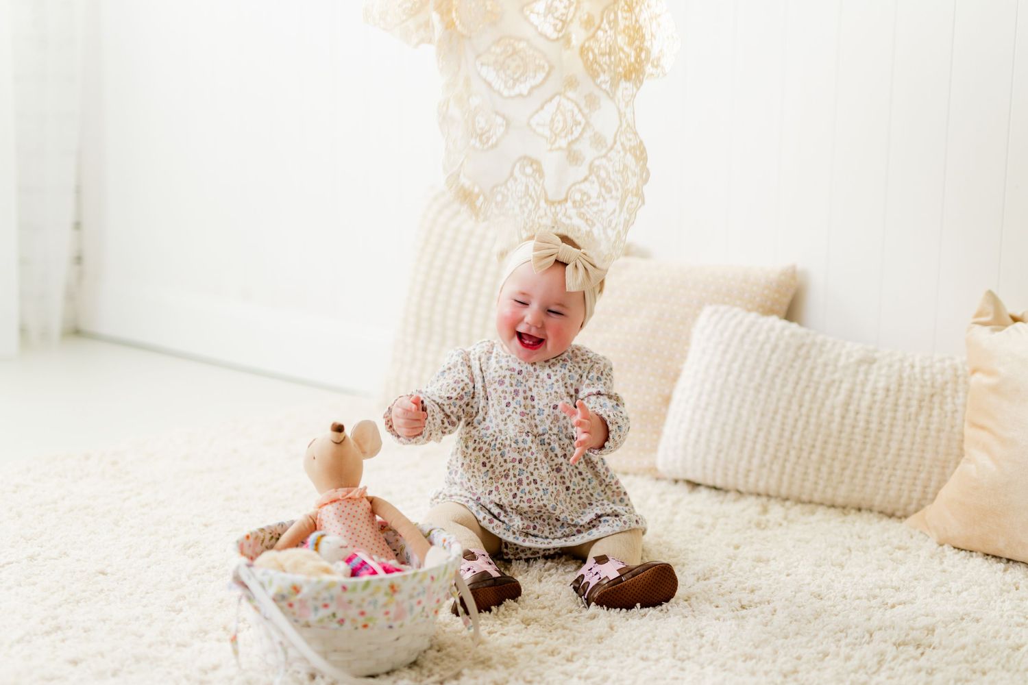 Happy baby girl in floral outfit sitting on white fluffy rug playing with toys and stuffed animal.