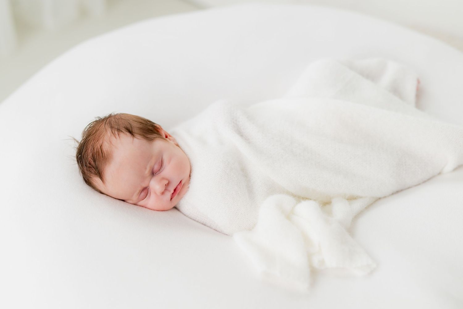 Peaceful newborn baby swaddled in white blanket sleeping on a round white surface in a bright, airy setting.