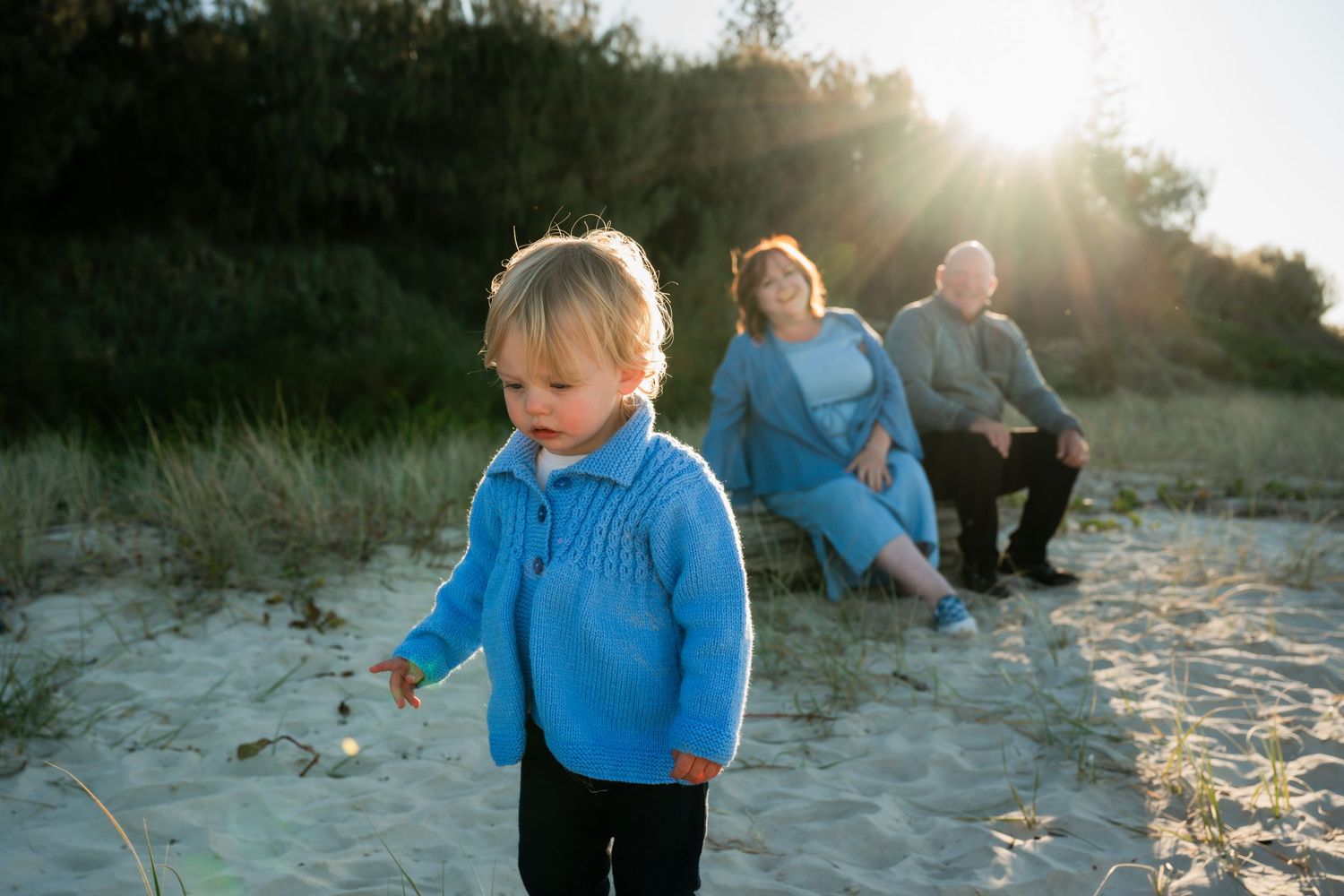 A toddler in a blue sweater plays on sandy beach dunes while grandparents sit together in warm sunset light.