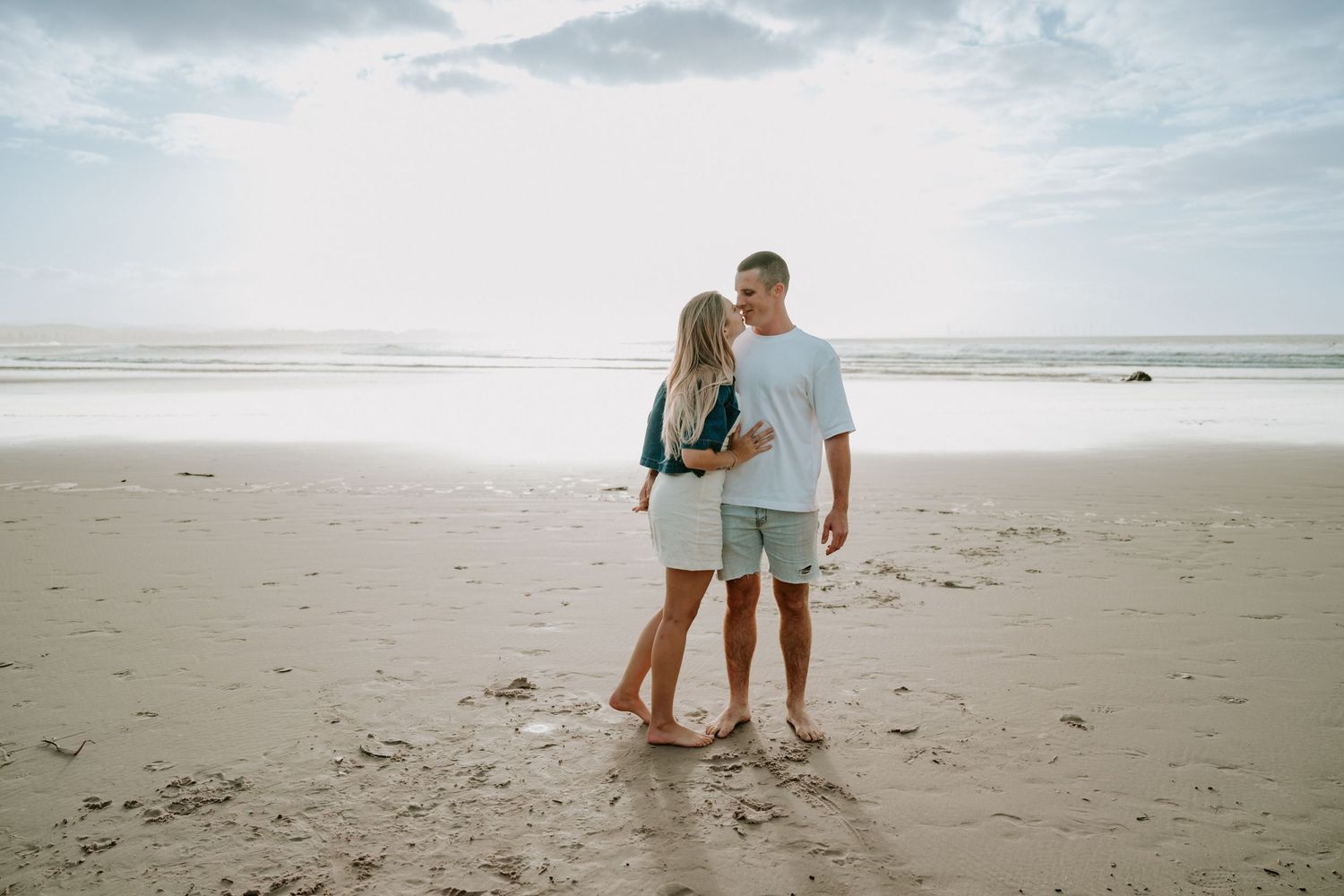 Couple embracing barefoot on a sandy beach with cloudy skies and ocean in the background.