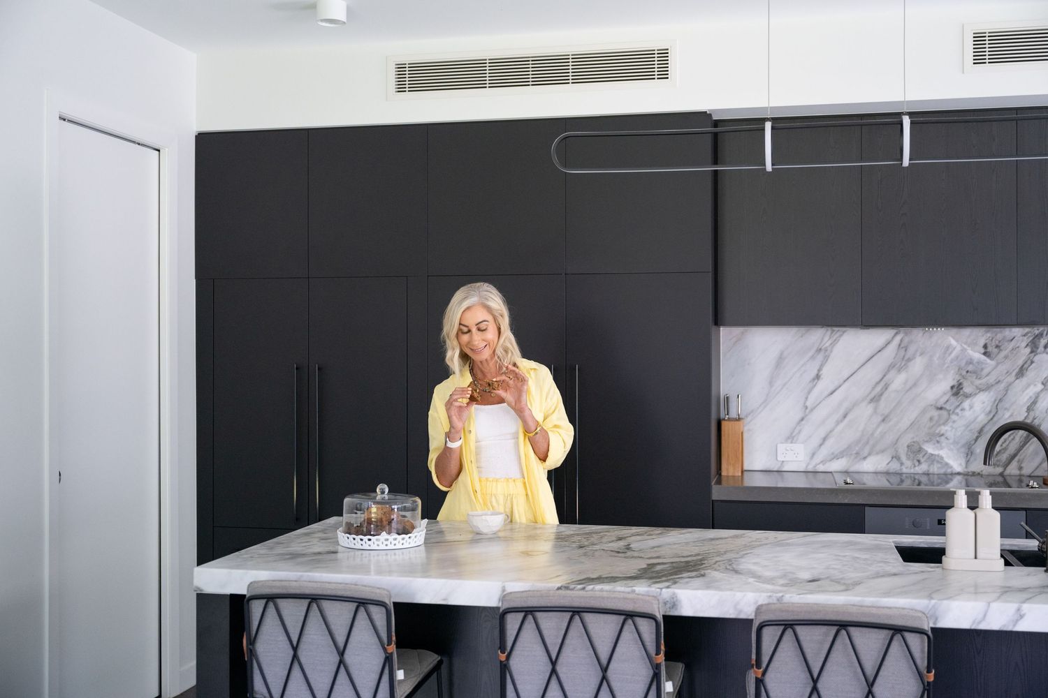 Older woman with gray hair in yellow jacket using phone at marble kitchen island with dark cabinets.