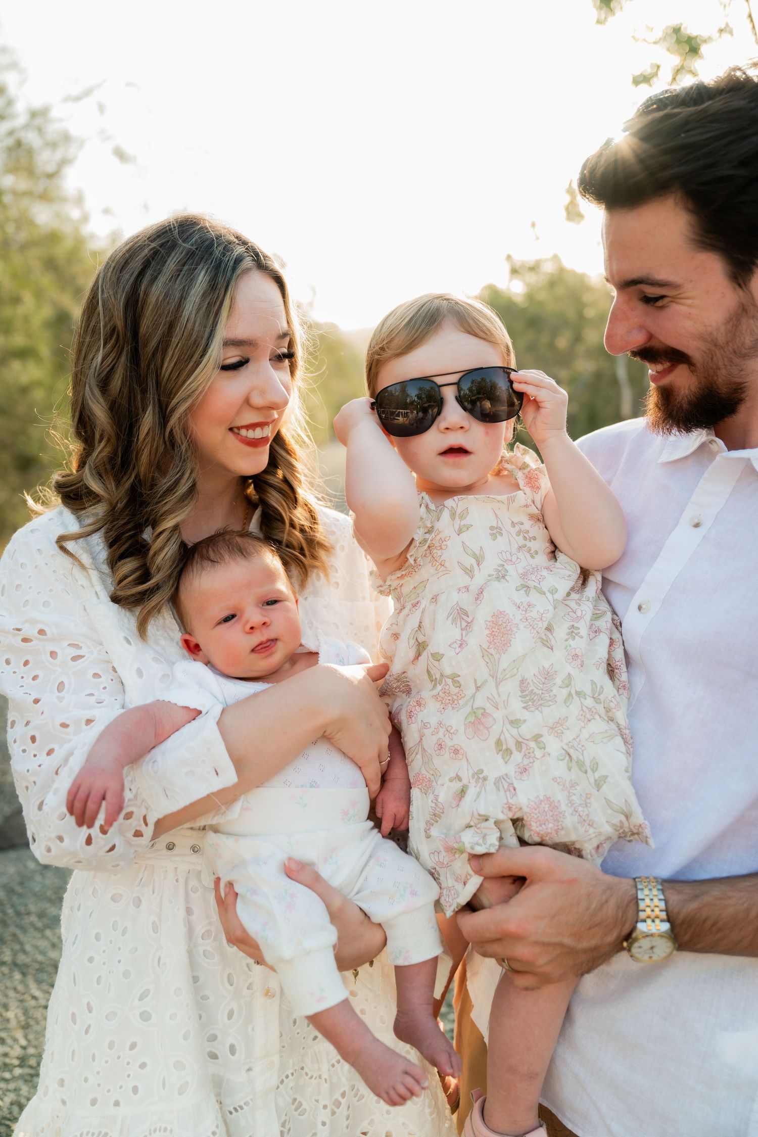 Young family portrait with mother in white lace dress holding newborn, father holding toddler girl wearing oversized sunglasses outdoors.
