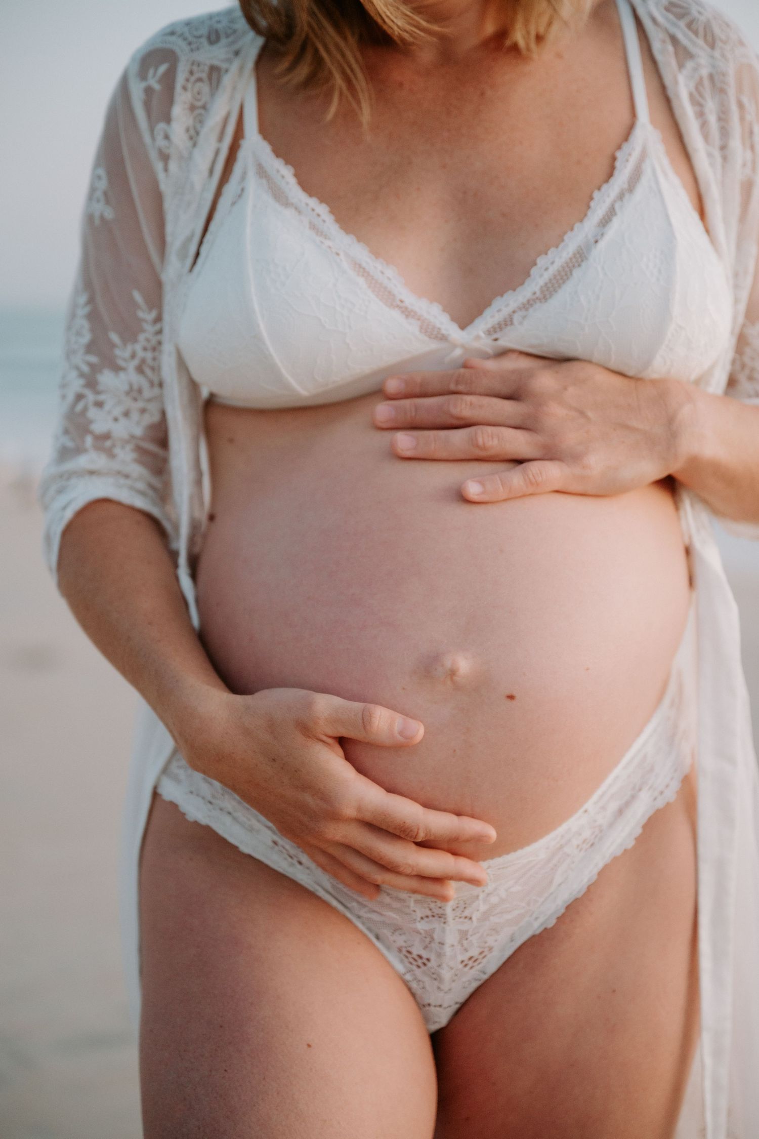 Pregnant woman in white lace lingerie cradling her baby bump on a beach.