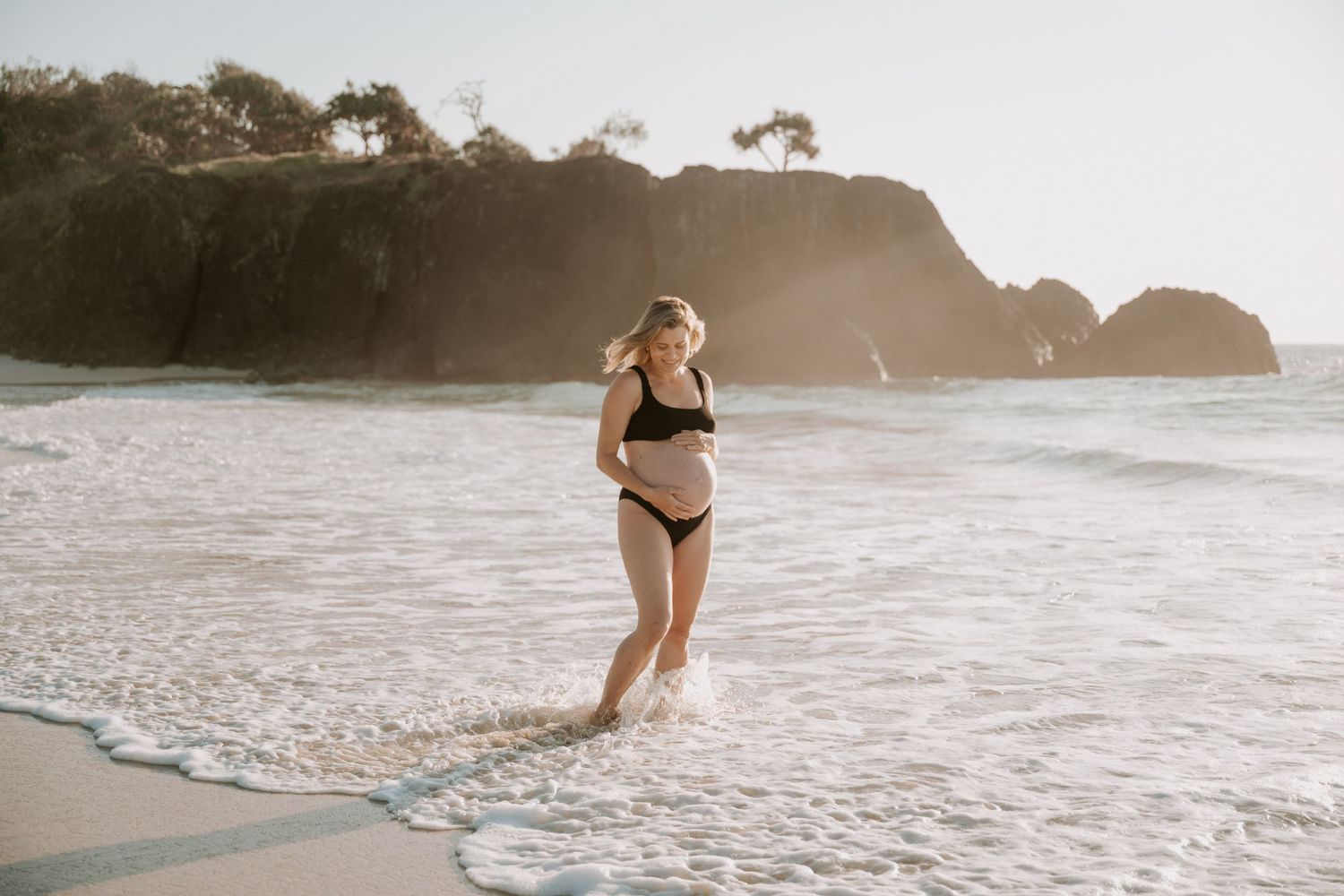 Pregnant woman in black bikini walking in ocean waves on a sunny beach with rocky cliffs in background.