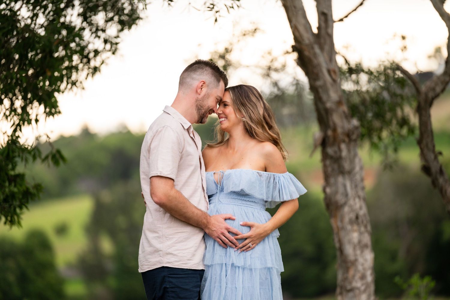Couple in maternity photoshoot outdoors, woman in blue dress cradling baby bump, man embracing her, lush green background.