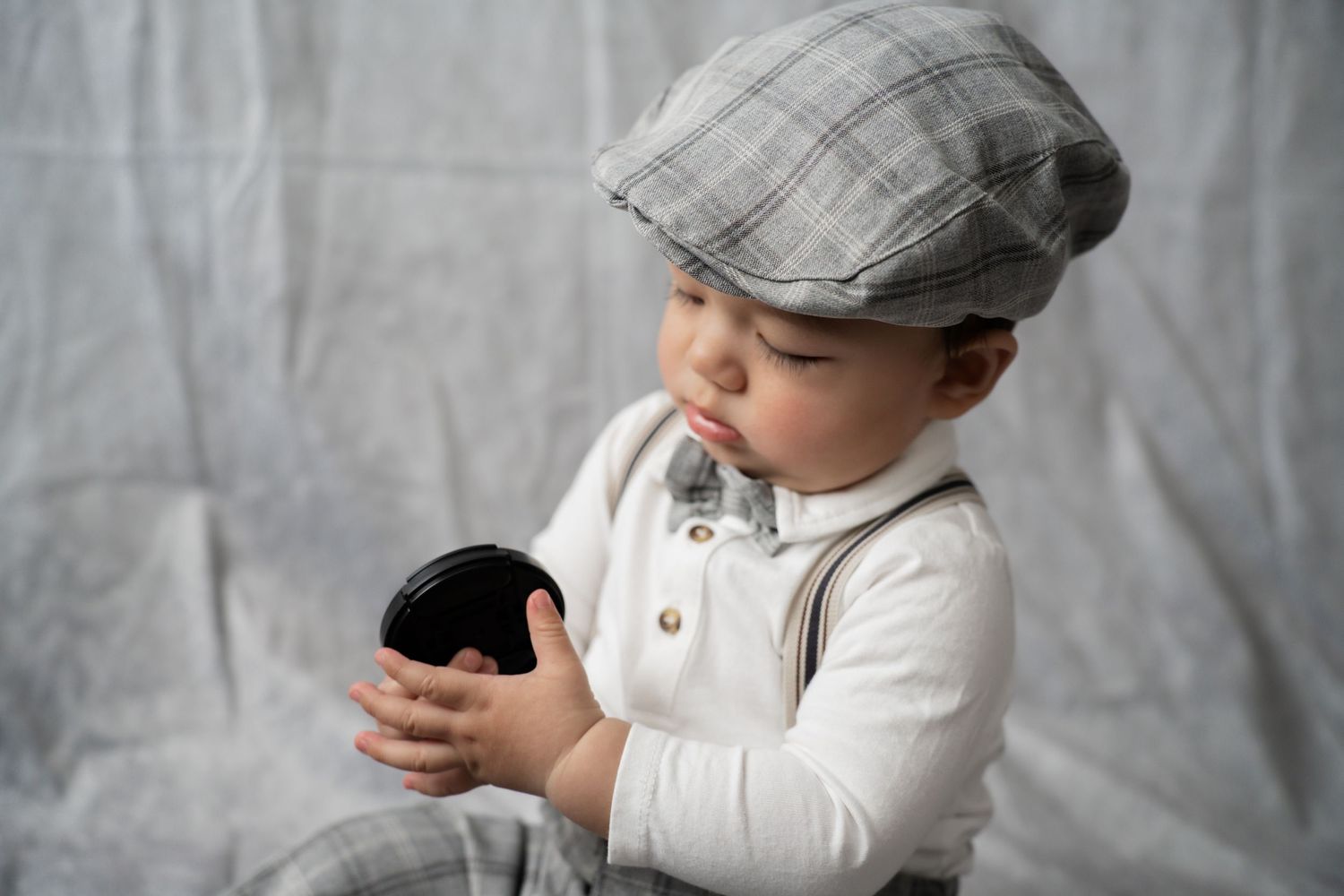 A stylish baby boy wearing a plaid flat cap, suspenders, and bow tie holds a dark object in a studio setting.
